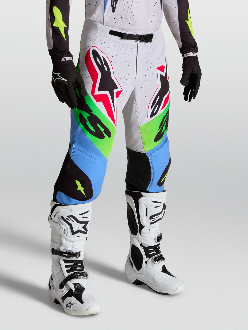 Alpinestars Spodnie Supertech Dyna, profesjonalne spodnie motocrossowe, zielone i niebieskie z białymi panelami, sportowy krój wyścigowy, na modelu z białymi butami i czarnymi rękawicami