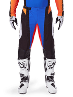 Alpinestars 2025 Techstar Melt Motocross Pants, pomarańczowo-niebieskie, noszone z pasującą koszulką i białymi butami, czarne panele z intensywnymi niebieskimi i pomarańczowymi akcentami, perforowane dla wentylacji, sportowy krój wyścigowy zapewniający wydajność w terenie.