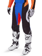 Alpinestars Techstar Melt Pants 2025, spodnie motocrossowe, czarne z niebieskimi i pomarańczowymi akcentami, noszone z białymi butami i dopasowaną koszulką, wykonane z wytrzymałego materiału ochronnego o sportowym, wyścigowym designie