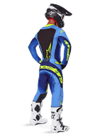 Spodnie Alpinestars Techstar Melt 2025, odzież motocrossowa, żółto-niebieskie (Yellow Fluo Blue), widok z tyłu na motocyklistę w dopasowanej niebieskiej koszulce z dużym żółtym logo, biało-czarne buty i czarny kask, skoordynowany strój wyścigowy do jazdy off-road