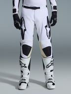 Alpinestars Racer Riway 2026, spodnie motocrossowe, biało-czarne, noszone z pasującymi biało-czarnymi butami motocrossowymi i czarnymi rękawicami, z wyrazistym czarnym logo Alpinestars na udach, techniczna konstrukcja do jazdy off-road
