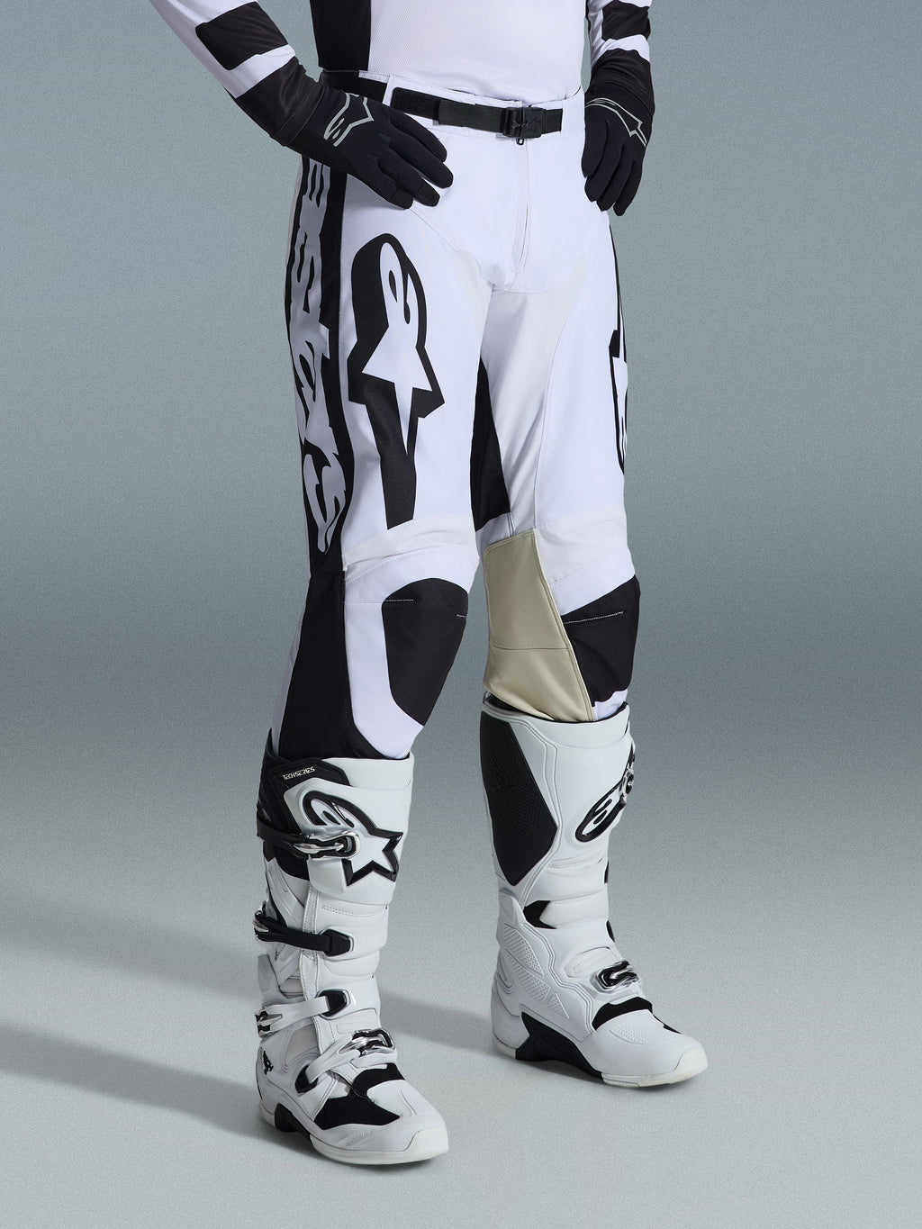 Alpinestars 2026 Racer Riway Pants, spodnie motocrossowe, biało-czarne, z wyraźną czarną grafiką i logo Alpinestars, beżowe skórzane wzmocnienia po wewnętrznej stronie kolan, noszone z biało-czarnymi butami motocrossowymi i czarnymi rękawicami, profesjonalna odzież wyścigowa do jazdy off-road.