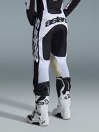 Alpinestars 2026 Racer Riway Pants, spodnie motocrossowe, biało-czarne, widok z tyłu ukazujący wyraźne czarne oznaczenia marki na siedzeniu i bokach, panele wewnętrzne kolan z beżowej koziej skóry, zestawione z pasującymi biało-czarnymi butami motocrossowymi