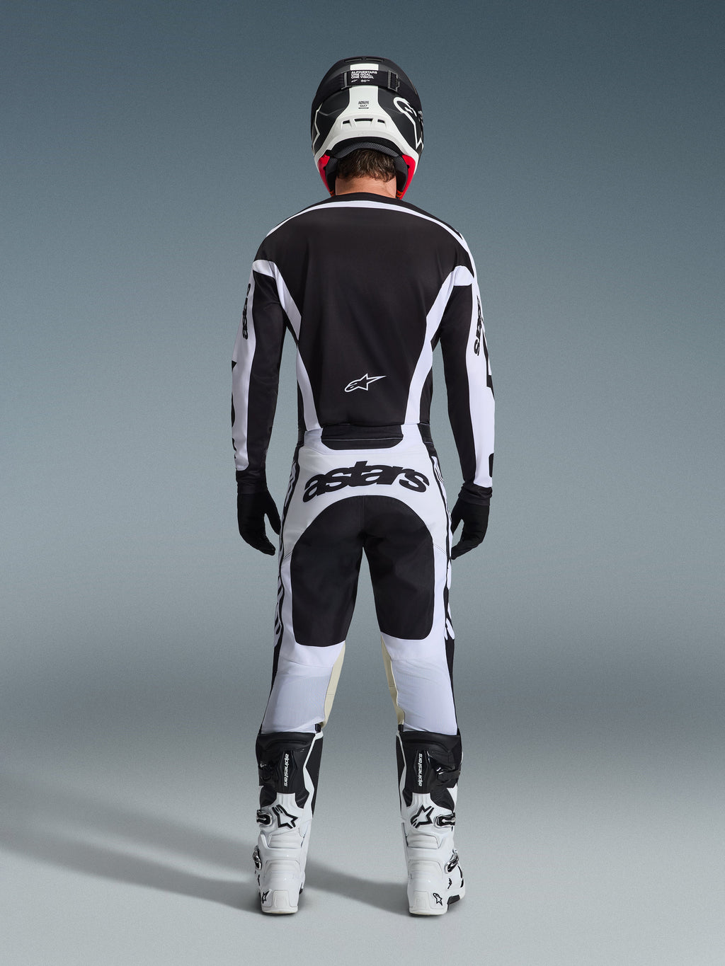 Alpinestars 2026 Racer Riway Pants, spodnie motocrossowe, biało-czarne, widok z tyłu na kompletny zestaw wyścigowy, wyraźny napis „astars” na siedzeniu, noszone z pasującą koszulką, białymi butami i czarnym kaskiem, dopasowany strój do wyścigów off-road.
