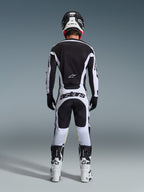 Alpinestars 2026 Racer Riway Pants, spodnie motocrossowe, biało-czarne, widok z tyłu na kompletny zestaw wyścigowy, wyraźny napis „astars” na siedzeniu, noszone z pasującą koszulką, białymi butami i czarnym kaskiem, dopasowany strój do wyścigów off-road.