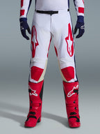 Spodnie Alpinestars Racer Riway 2026, spodnie motocrossowe, biało-niebiesko-czerwone, zestawione z czerwonymi butami i granatowymi rękawicami, duże czerwone logo na udach i czerwone panele na kolanach, sportowy, profilowany krój z regulowaną klamrą w pasie