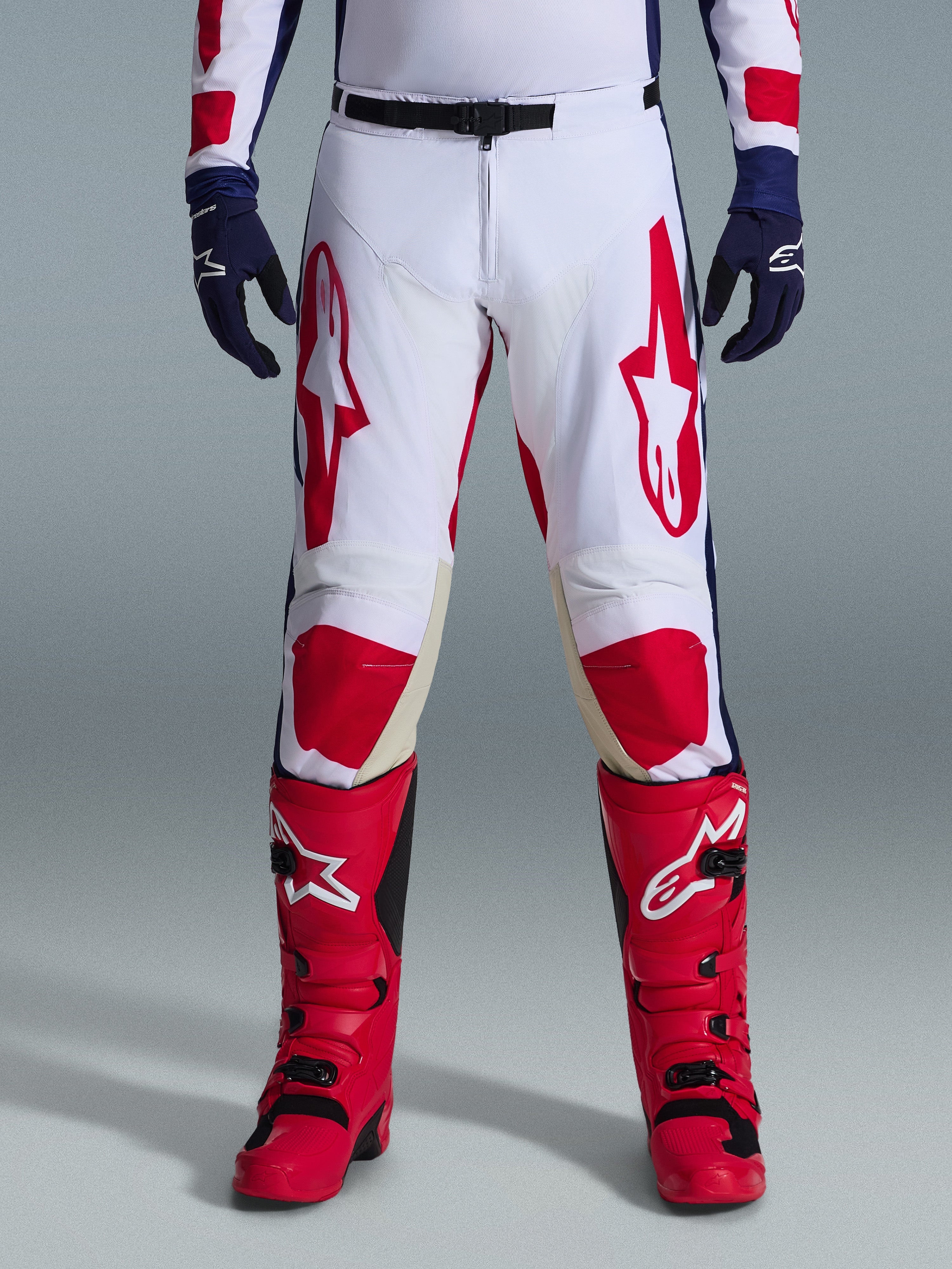 Spodnie Alpinestars Racer Riway 2026, spodnie motocrossowe, biało-niebiesko-czerwone, zestawione z czerwonymi butami i granatowymi rękawicami, duże czerwone logo na udach i czerwone panele na kolanach, sportowy, profilowany krój z regulowaną klamrą w pasie