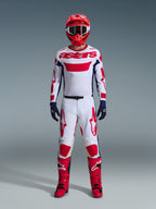 Motocyklista motocrossowy w kompletnym stroju wyścigowym, biała koszulka z czerwonym brandingiem Alpinestars i niebieskimi akcentami, spodnie Alpinestars 2026 Racer Riway w kolorystyce White Blue Red, czerwony kask z dopasowanymi goglami, czerwono-czarne buty, granatowe rękawice, skoordynowany zestaw do wyścigów terenowych