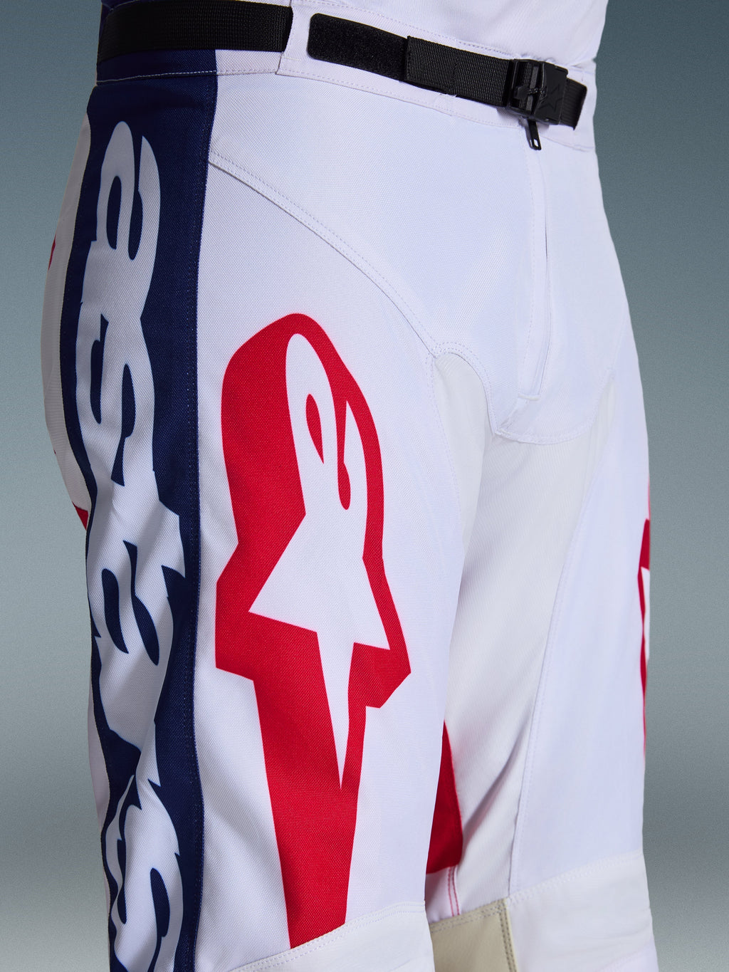 Alpinestars Racer Riway Pants 2026, spodnie motocrossowe, biało-niebiesko-czerwone, zbliżenie na górną część nogawki i talię pokazujące duże czerwone logo gwiazdy na białym materiale poliestrowym, niebieskie panele boczne z brandingiem oraz czarny regulowany pasek w pasie z magnetyczną klamrą SX2, sportowy design wyścigowy off-road