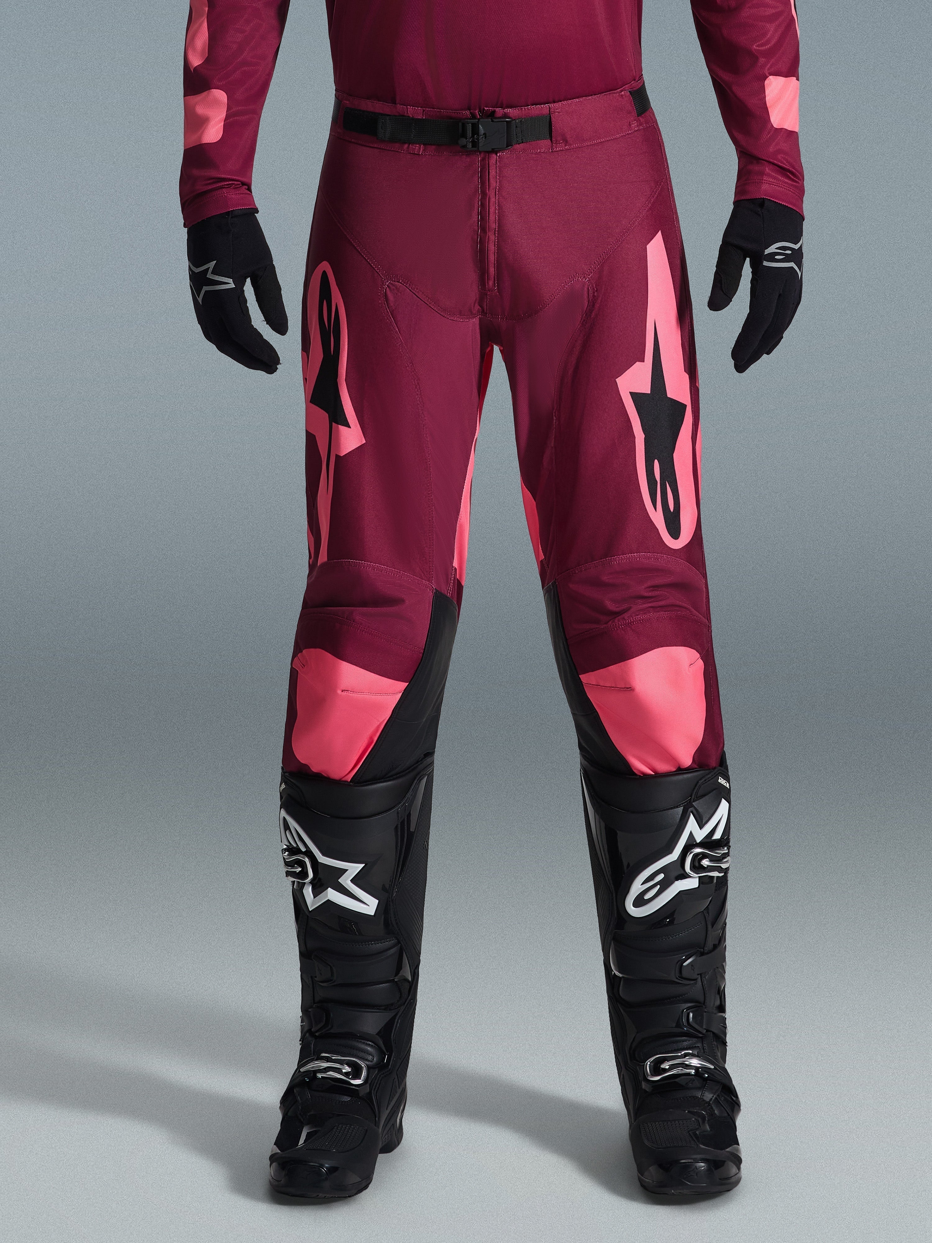 Alpinestars Racer Riway Pants 2026, spodnie motocrossowe, ciemna czerwień koralowa, noszone z czarnymi rękawicami i butami, duże koralowe logo Alpinestars na udach, czarna klamra w pasie, wstępnie zakrzywiona konstrukcja nogawek do jazdy terenowej