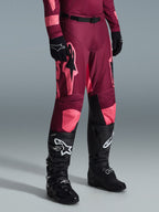 Alpinestars 2026 Racer Riway Pants, spodnie motocrossowe, kolor Dark Red Coral, z dużymi różowymi logotypami gwiazdy i czarnymi panelami po wewnętrznej stronie kolan, noszone z czarnymi butami i rękawicami, sportowy krój wyścigowy do jazdy w terenie