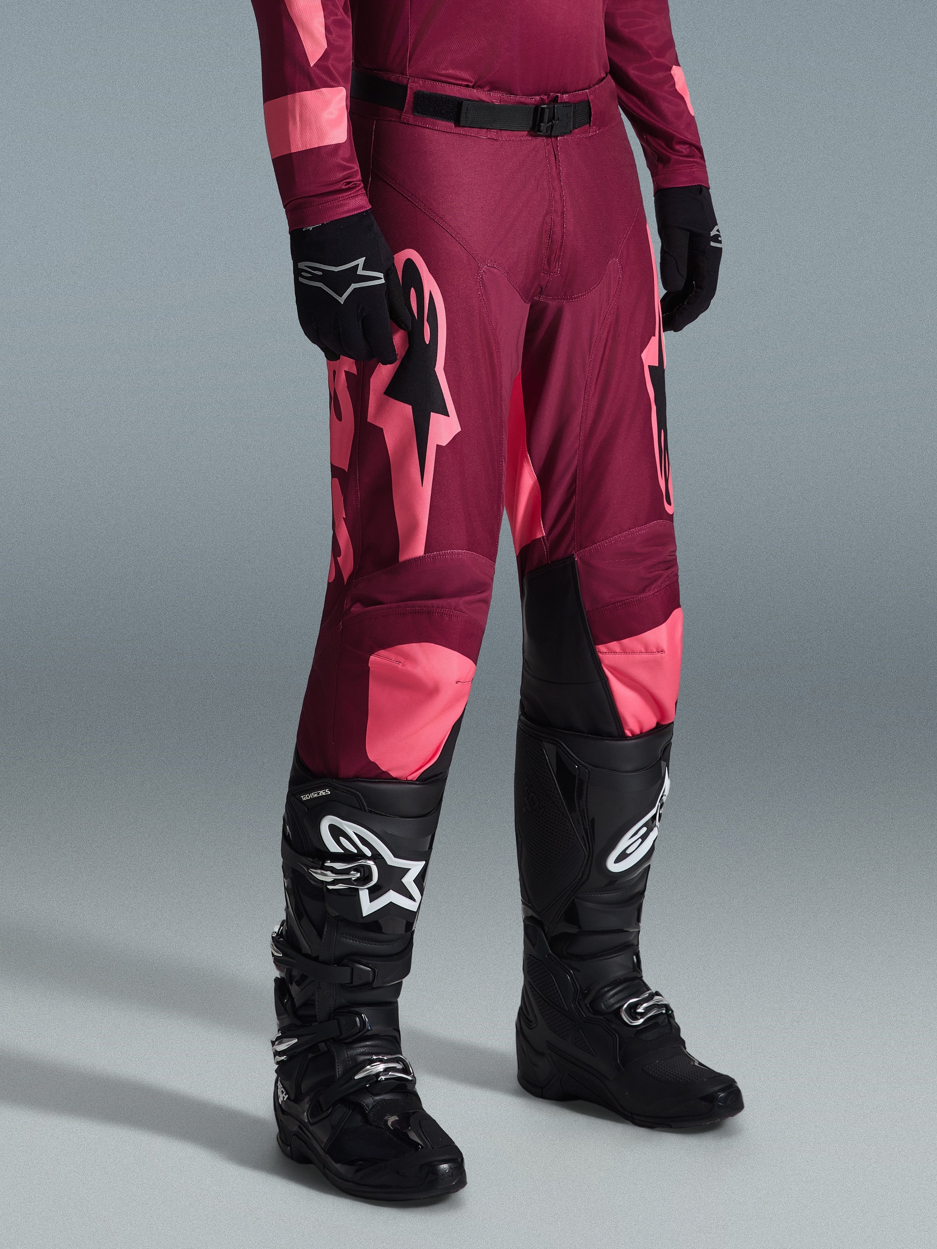 Alpinestars 2026 Racer Riway Pants, spodnie motocrossowe, kolor Dark Red Coral, z dużymi różowymi logotypami gwiazdy i czarnymi panelami po wewnętrznej stronie kolan, noszone z czarnymi butami i rękawicami, sportowy krój wyścigowy do jazdy w terenie