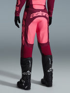 Alpinestars Racer Riway Pants 2026, spodnie motocrossowe, ciemnoczerwone i koralowe, widok z tyłu pokazujący duże koralowe logo „astars” na siedzeniu, noszone z czarnymi butami i rękawicami motocrossowymi, odzież wyczynowa do jazdy off-road