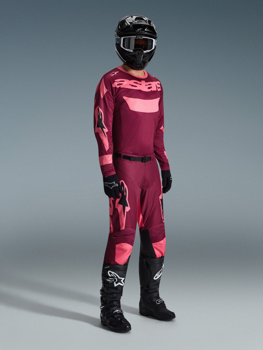 Alpinestars Racer Riway Pants 2026, spodnie motocrossowe, w kolorze Dark Red Coral, noszone z pasującą koszulką, czarnym kaskiem i czarnymi butami, z różowym logo Alpinestars i czarnymi panelami na kolanach, profesjonalna odzież do wyścigów off-road