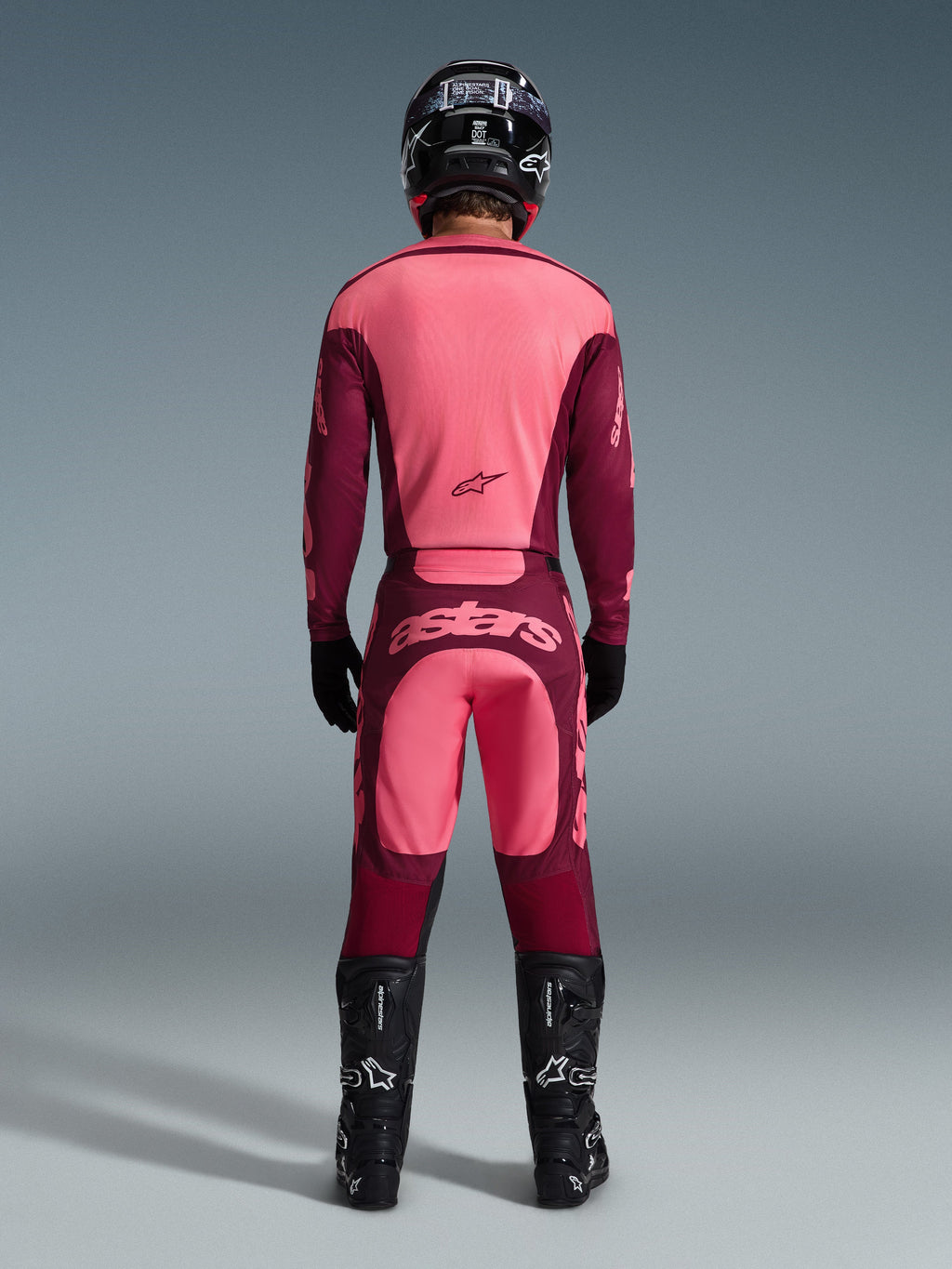 Zawodnik motocrossowy widziany od tyłu w koszulce Alpinestars 2026 Racer Riway i pasujących spodniach w kolorze Dark Red Coral, czarny kask, czarne rękawice i czarne buty, profesjonalny zestaw odzieży do jazdy terenowej
