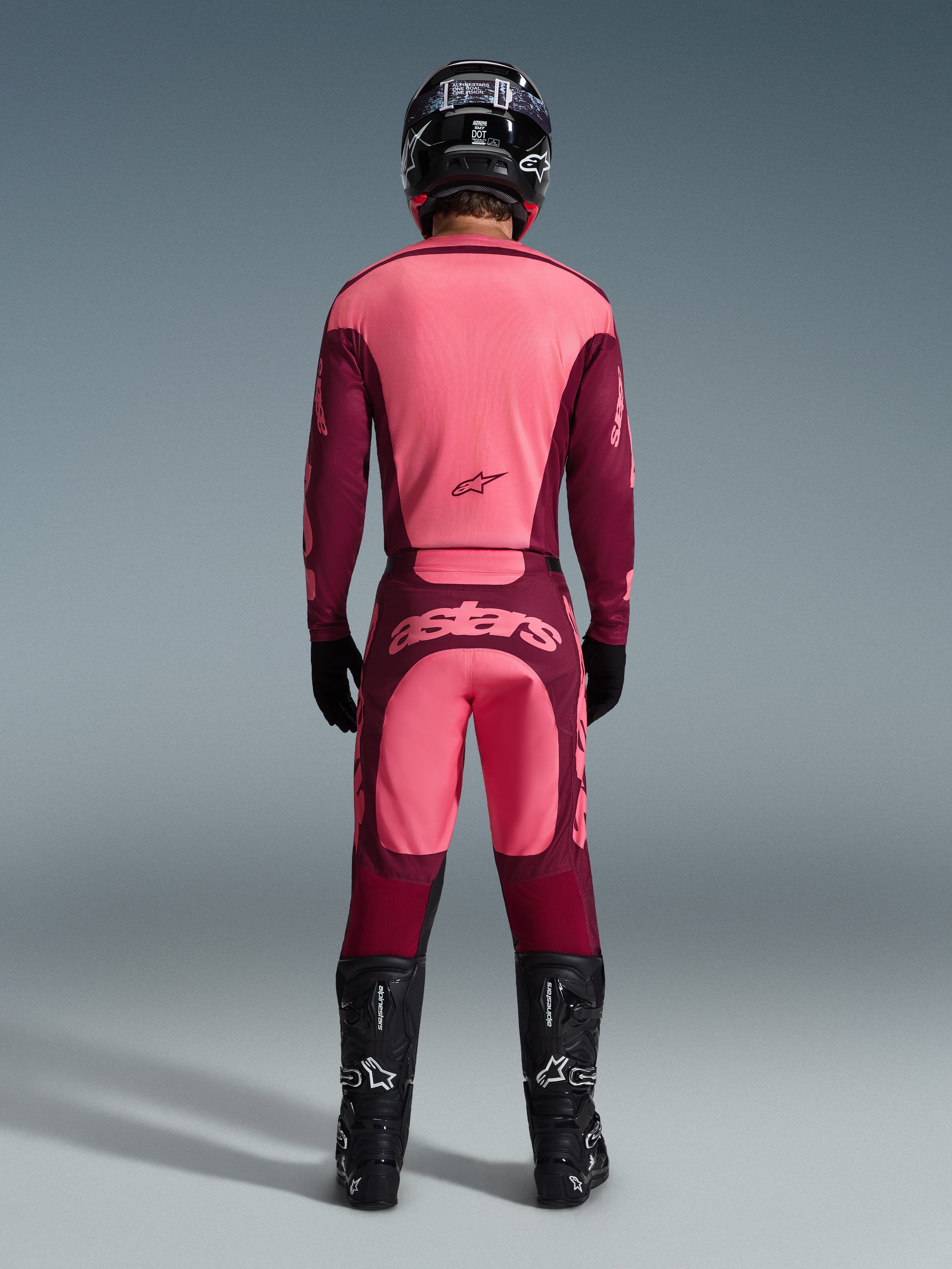 Zawodnik motocrossowy widziany od tyłu w koszulce Alpinestars 2026 Racer Riway i pasujących spodniach w kolorze Dark Red Coral, czarny kask, czarne rękawice i czarne buty, profesjonalny zestaw odzieży do jazdy terenowej