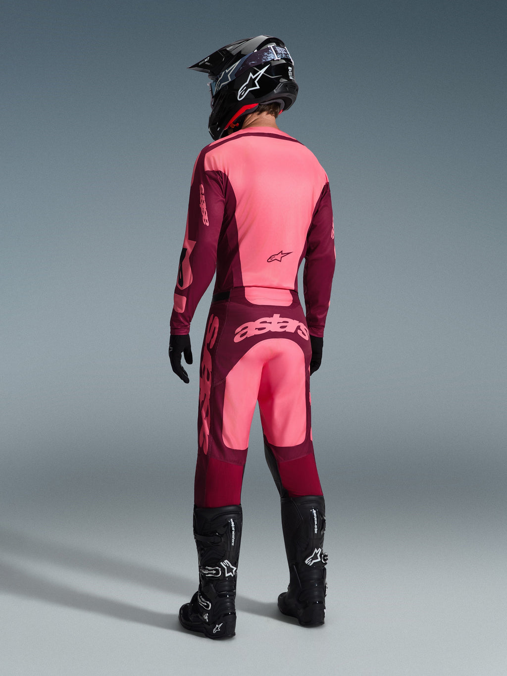 Spodnie Alpinestars Racer Riway 2026, spodnie motocrossowe, kolor Dark Red Coral, widok z tyłu na motocyklistę w pasującej koszulce, czarnym kasku i czarnych butach, z wyraźnym koralowym logo na siedzeniu i ergonomicznym sportowym krojem do jazdy off-road