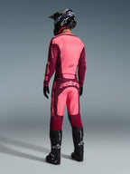 Spodnie Alpinestars Racer Riway 2026, spodnie motocrossowe, kolor Dark Red Coral, widok z tyłu na motocyklistę w pasującej koszulce, czarnym kasku i czarnych butach, z wyraźnym koralowym logo na siedzeniu i ergonomicznym sportowym krojem do jazdy off-road