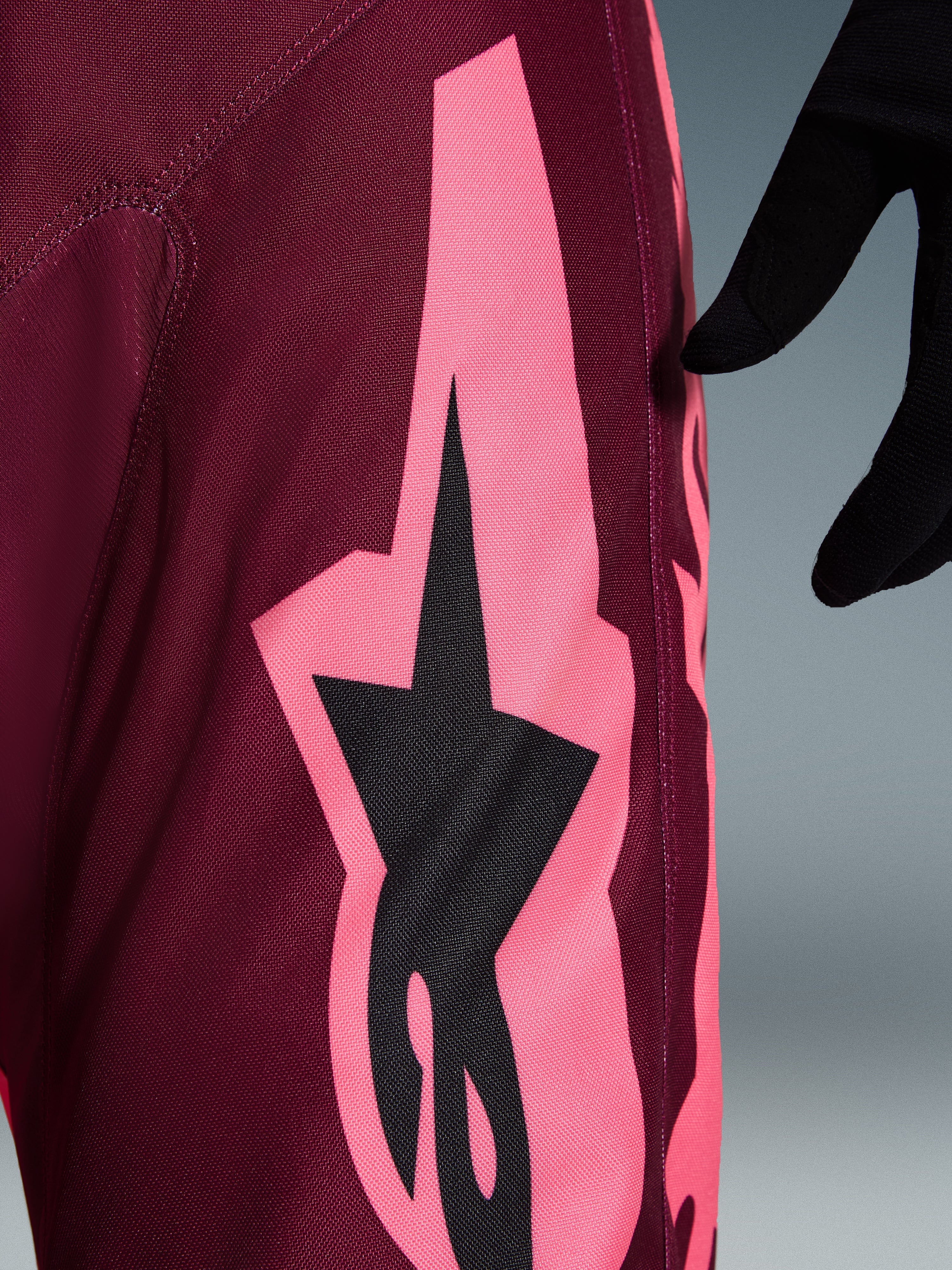 Alpinestars 2026 Racer Riway Pants, spodnie motocrossowe, kolor Dark Red Coral, widok z bliska na dużą koralowo-różową kanciastą grafikę i czarne logo gwiazdy, podkreślający wytrzymałą strukturę materiału polyfabric i sportowe szwy