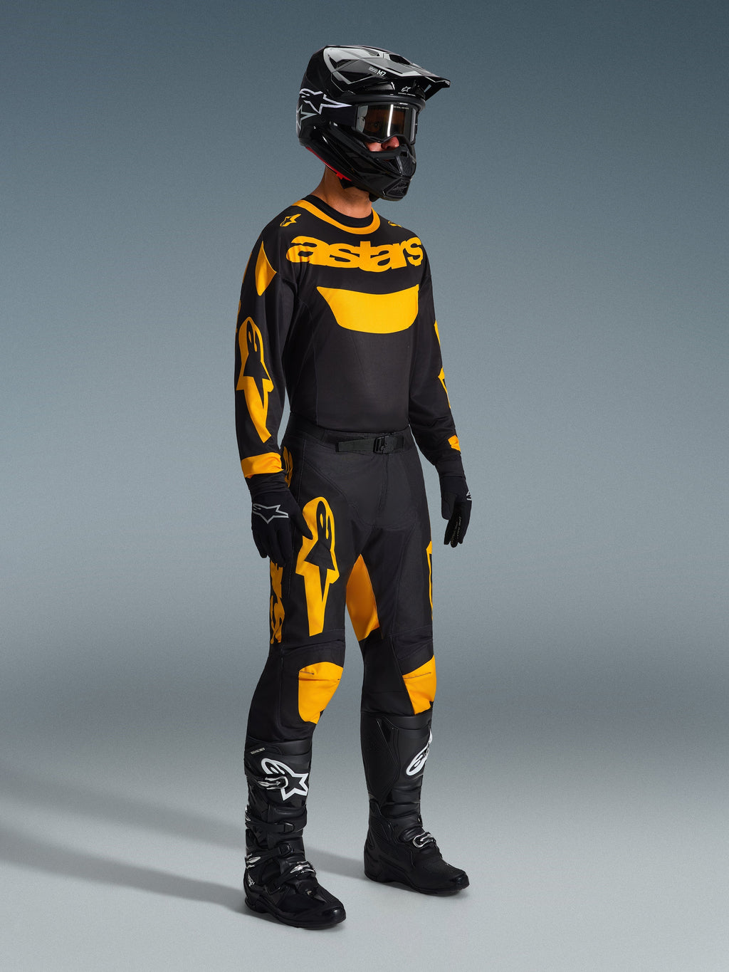 Motocyklista motocrossowy ubrany w spodnie Alpinestars 2026 Racer Riway w kolorze żółto-czarnym, w zestawieniu z pasującą czarno-żółtą koszulką „astars”, czarnym kaskiem z goglami i czarnymi butami wyścigowymi; skoordynowany zestaw odzieży do jazdy motocrossowej off-road.