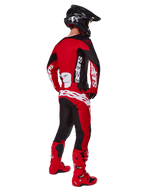 Zestaw Alpinestars 2025 Racer Veil, motocyklista motocrossowy widziany od tyłu w czerwonej koszulce z czarnymi panelami i białym logo, pasujące czerwono-czarne spodnie z dużymi białymi oznaczeniami marki, czerwone buty i czarny kask, kompletny strój wyścigowy off-road