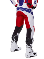 Alpinestars 2025 Racer Veil Pants, spodnie motocrossowe, jasnoczerwone i białe z niebieskim brandingiem, widok z tyłu ukazujący czarne elastyczne panele wentylacyjne z tyłu nogawek, zestawione z białymi butami motocrossowymi.