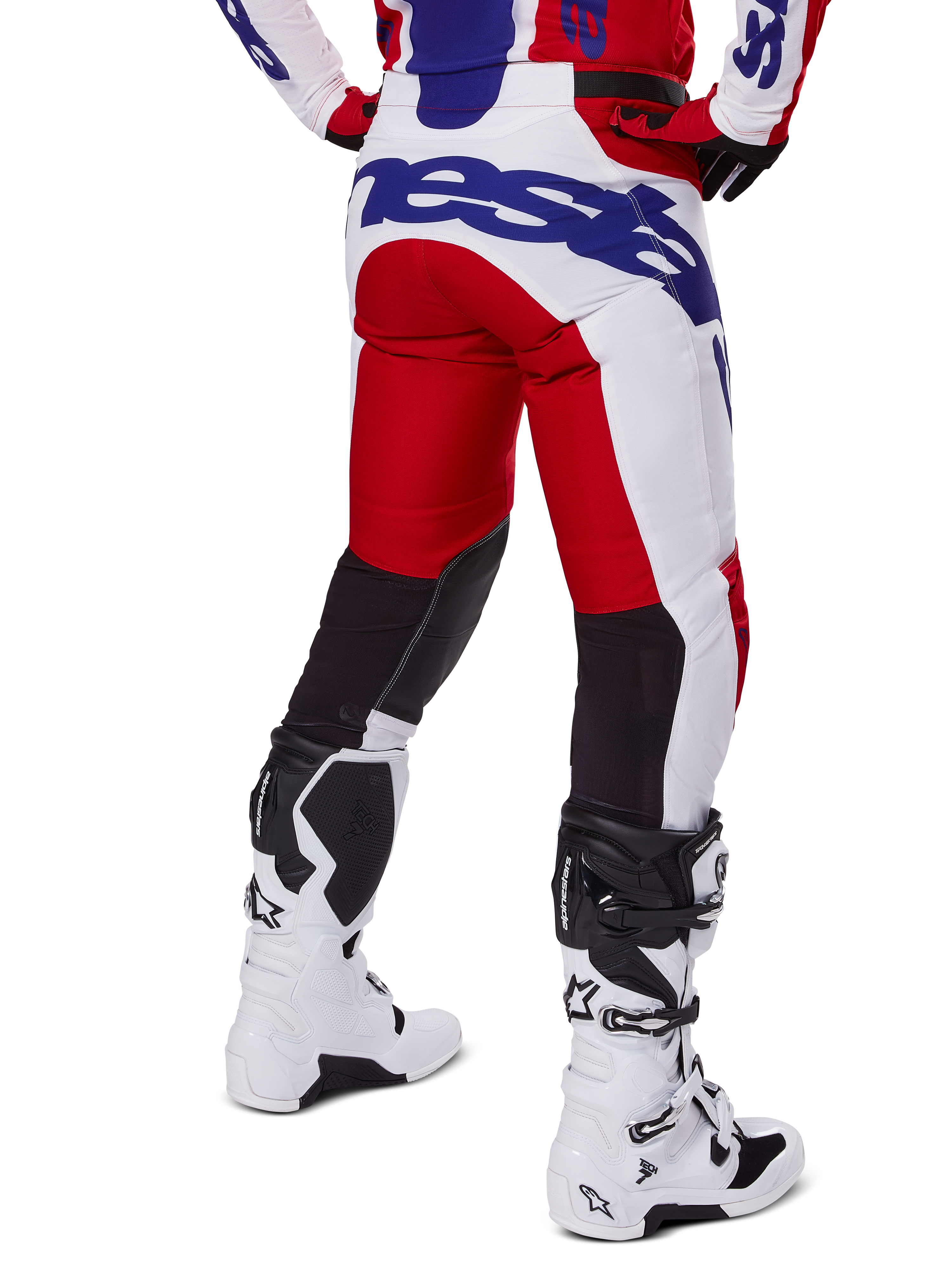 Alpinestars 2025 Racer Veil Pants, spodnie motocrossowe, jasnoczerwone i białe z niebieskim brandingiem, widok z tyłu ukazujący czarne elastyczne panele wentylacyjne z tyłu nogawek, zestawione z białymi butami motocrossowymi.