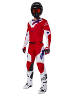 Spodnie Alpinestars Racer Veil 2025, spodnie motocrossowe, jaskrawoczerwone i białe, prezentowane jako część pełnego zestawu wyścigowego z dopasowaną koszulką, czarnym kaskiem z pomarańczowymi goglami, białymi butami i czerwonymi rękawicami, z wyraźnym niebieskim logo Alpinestars.
