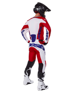 Alpinestars Racer Veil Pants 2025, spodnie motocrossowe, jaskrawoczerwone i białe z niebieskimi akcentami, widok od tyłu na motocyklistę w pełnym stroju, wyraźne logo Alpinestars, oddychające panele na dolnej części nogawek, białe buty i czarny kask