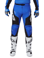 Alpinestars Racer Veil Pants 2025, spodnie motocrossowe, niebiesko-czarne z białym logo Alpinestars na nogawkach, czarne panele boczne, noszone z białymi butami motocrossowymi i niebiesko-szarymi rękawicami, wysokowydajny sprzęt do jazdy terenowej