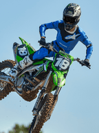 Zawodnik motocrossowy podczas skoku na zielonym motocyklu, ubrany w spodnie Alpinestars 2025 Racer Veil w kolorze niebiesko-czarnym, pasującą niebieską koszulkę z białym logo na piersi, biało-czarne buty i czarny kask, profesjonalna odzież wyścigowa off-road