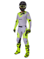 Alpinestars Racer Veil Pants 2025, spodnie motocrossowe, szare i żółte fluo, noszone z pasującą koszulką, czarnym kaskiem, żółtymi goglami fluo, butami i rękawicami, skoordynowany strój do wyścigów terenowych z wyrazistym brandingiem i ergonomicznym designem.