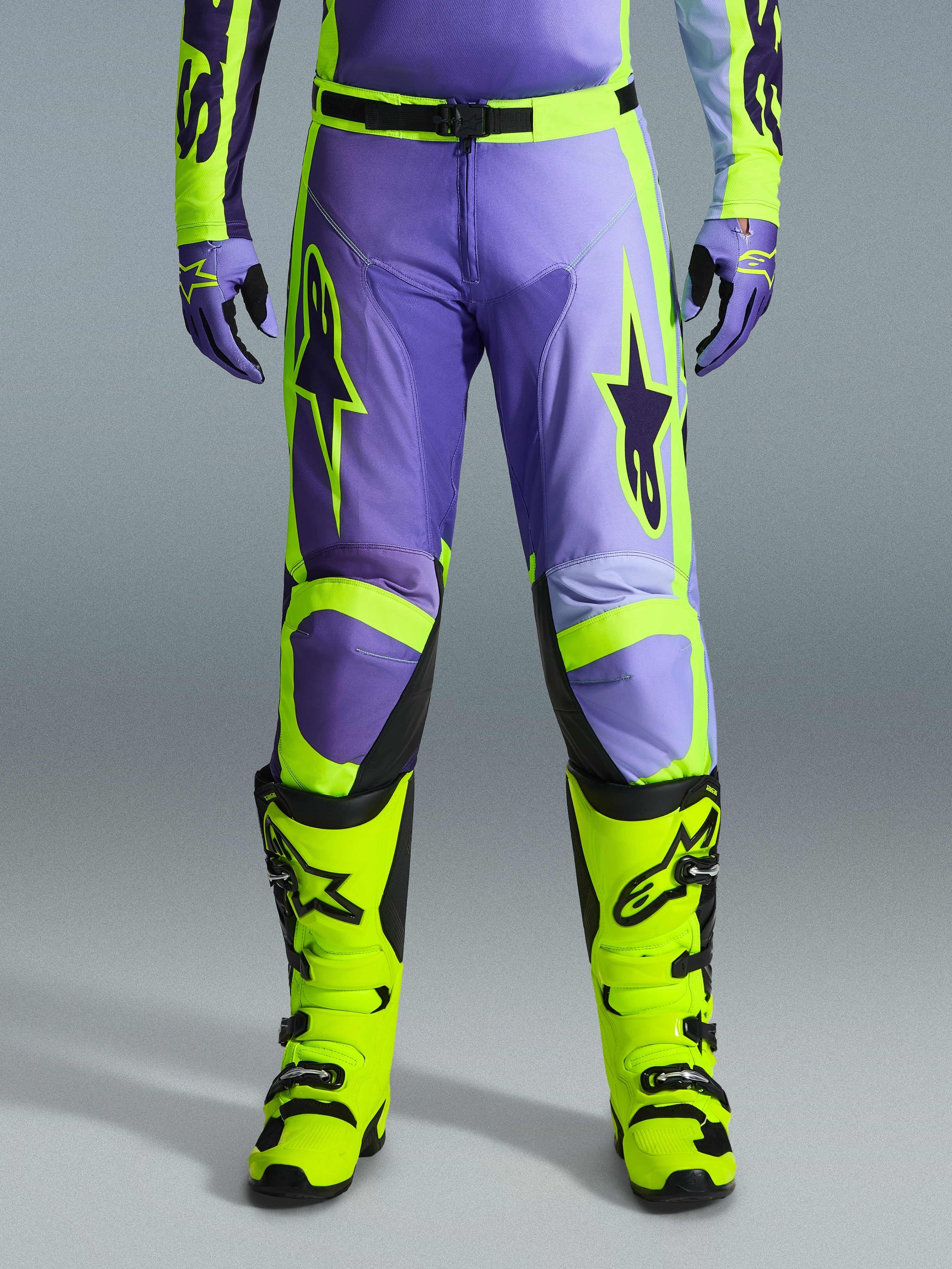 Alpinestars 2026 Racer Portl Pants, spodnie motocrossowe, fioletowe i żółte neonowe, noszone z dopasowanymi żółtymi neonowymi butami i fioletowymi rękawicami, z dużym logo gwiazdy i wytrzymałą konstrukcją z tkaniny poliestrowej do wyścigów terenowych