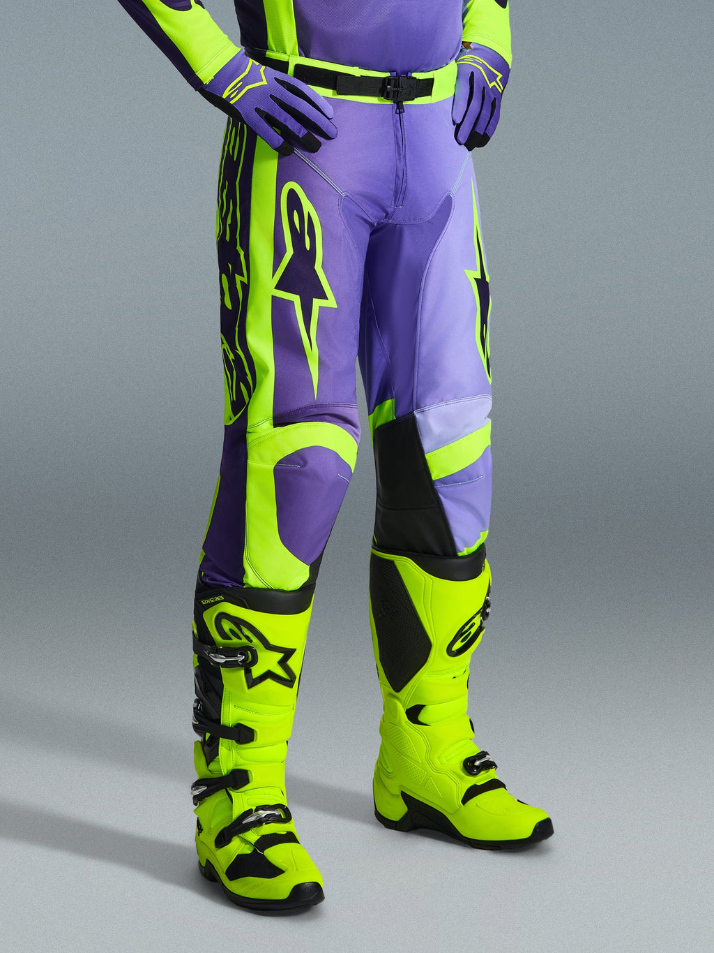 Alpinestars 2026 Racer Portl Pants, spodnie motocrossowe w kolorze fioletowym i żółtym fluo, noszone przez motocyklistę z pasującymi żółtymi neonowymi butami i fioletowymi rękawicami, z wyraźnym logo Alpinestars i ergonomiczną konstrukcją zapewniającą wydajność w terenie