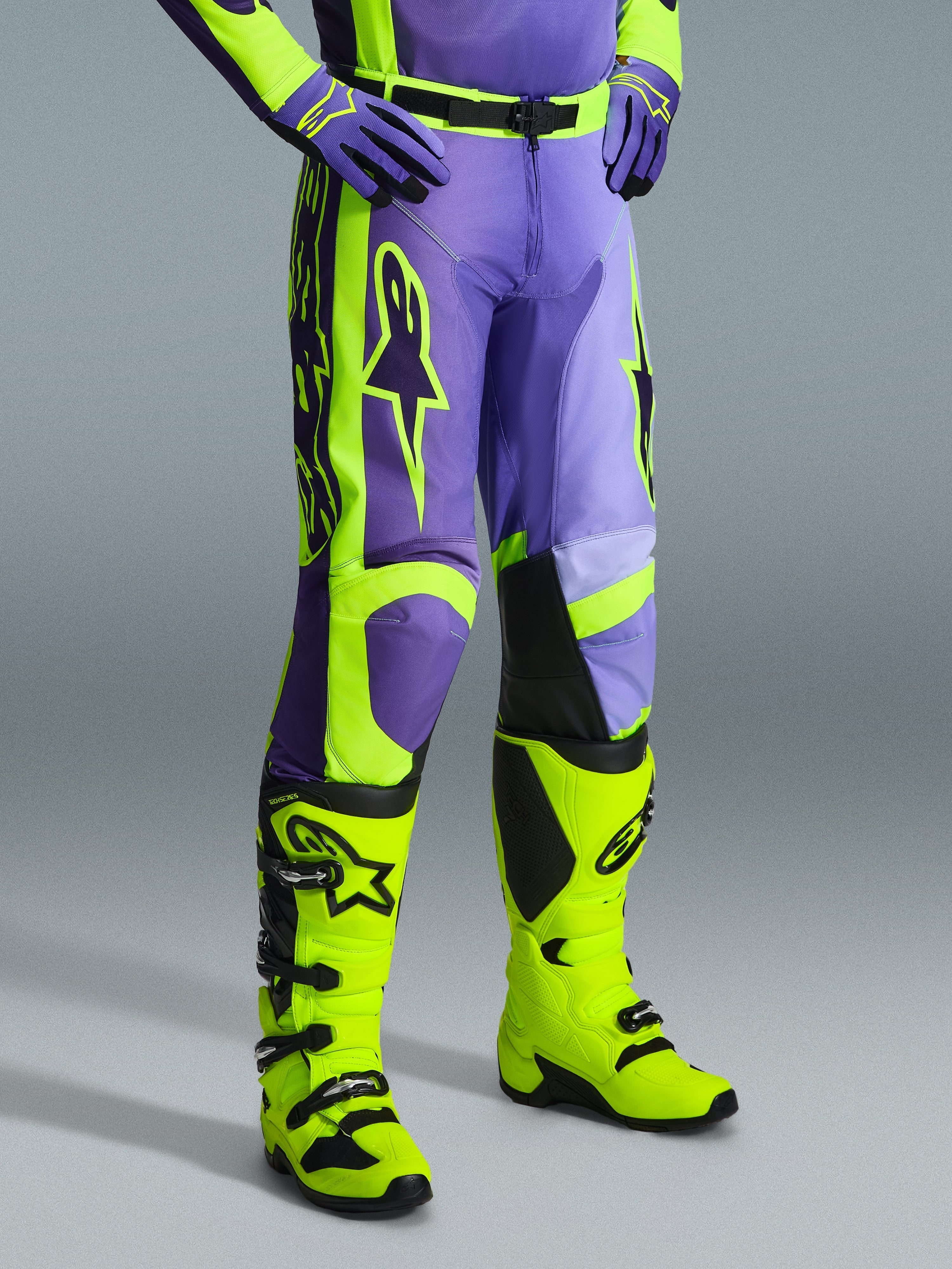 Alpinestars 2026 Racer Portl Pants, spodnie motocrossowe w kolorze fioletowym i żółtym fluo, noszone przez motocyklistę z pasującymi żółtymi neonowymi butami i fioletowymi rękawicami, z wyraźnym logo Alpinestars i ergonomiczną konstrukcją zapewniającą wydajność w terenie