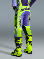 Alpinestars 2026 Racer Portl Pants, spodnie motocrossowe, fioletowe i żółte neonowe, widok z tyłu z wyraźnym brandingiem astars i fioletowymi akcentami, noszone z dopasowanymi żółtymi neonowymi butami motocrossowymi, wytrzymały materiał syntetyczny do wyścigów off-road