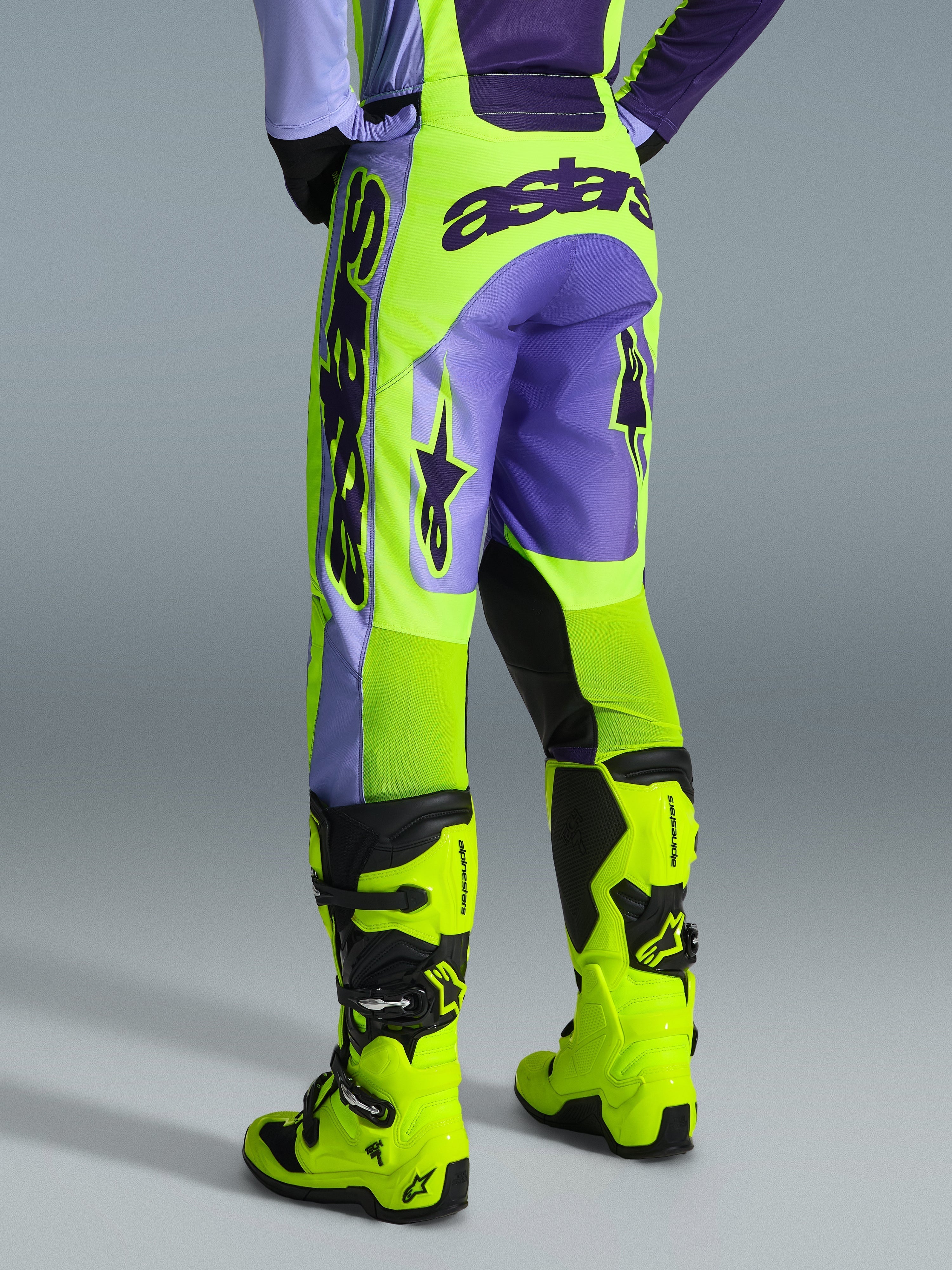 Alpinestars 2026 Racer Portl Pants, spodnie motocrossowe, fioletowe i żółte neonowe, widok z tyłu z wyraźnym brandingiem astars i fioletowymi akcentami, noszone z dopasowanymi żółtymi neonowymi butami motocrossowymi, wytrzymały materiał syntetyczny do wyścigów off-road