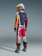 Zawodnik motocrossowy widziany od tyłu w pełnym stroju wyścigowym, spodnie Alpinestars 2026 Racer Portl w kolorach czerwonym, jasnoszarym i ciemnoszarym, dopasowana koszulka z pomarańczowymi i bordowymi rękawami, wielokolorowy kask z grafiką, szaro-czarne buty off-road z różowymi akcentami, profesjonalny zestaw motocrossowy