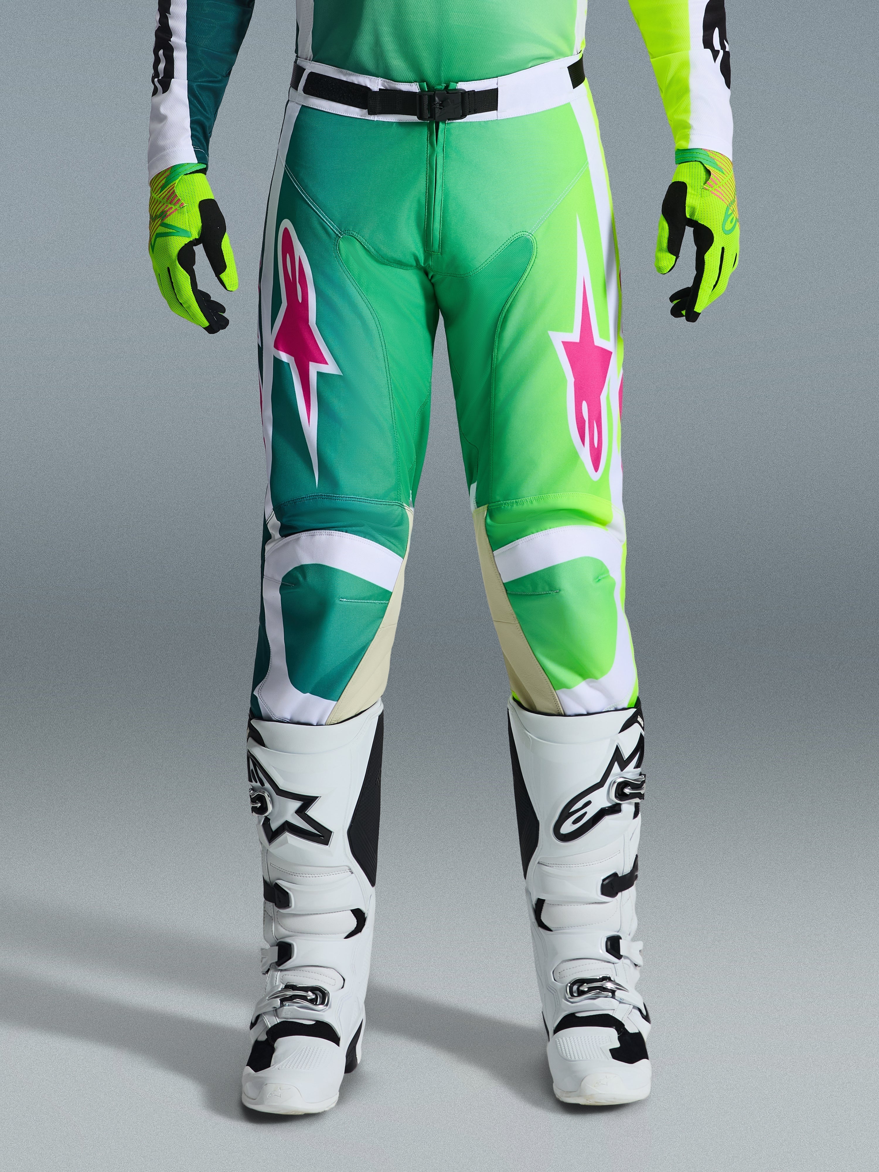 Alpinestars 2026 Racer Portl Pants, spodnie motocrossowe, zielone, białe i czarne z różowymi logotypami gwiazdy, noszone z biało-czarnymi butami i limonkowo-zielonymi rękawicami, gradientowy design i ergonomiczny krój do wyścigów off-road