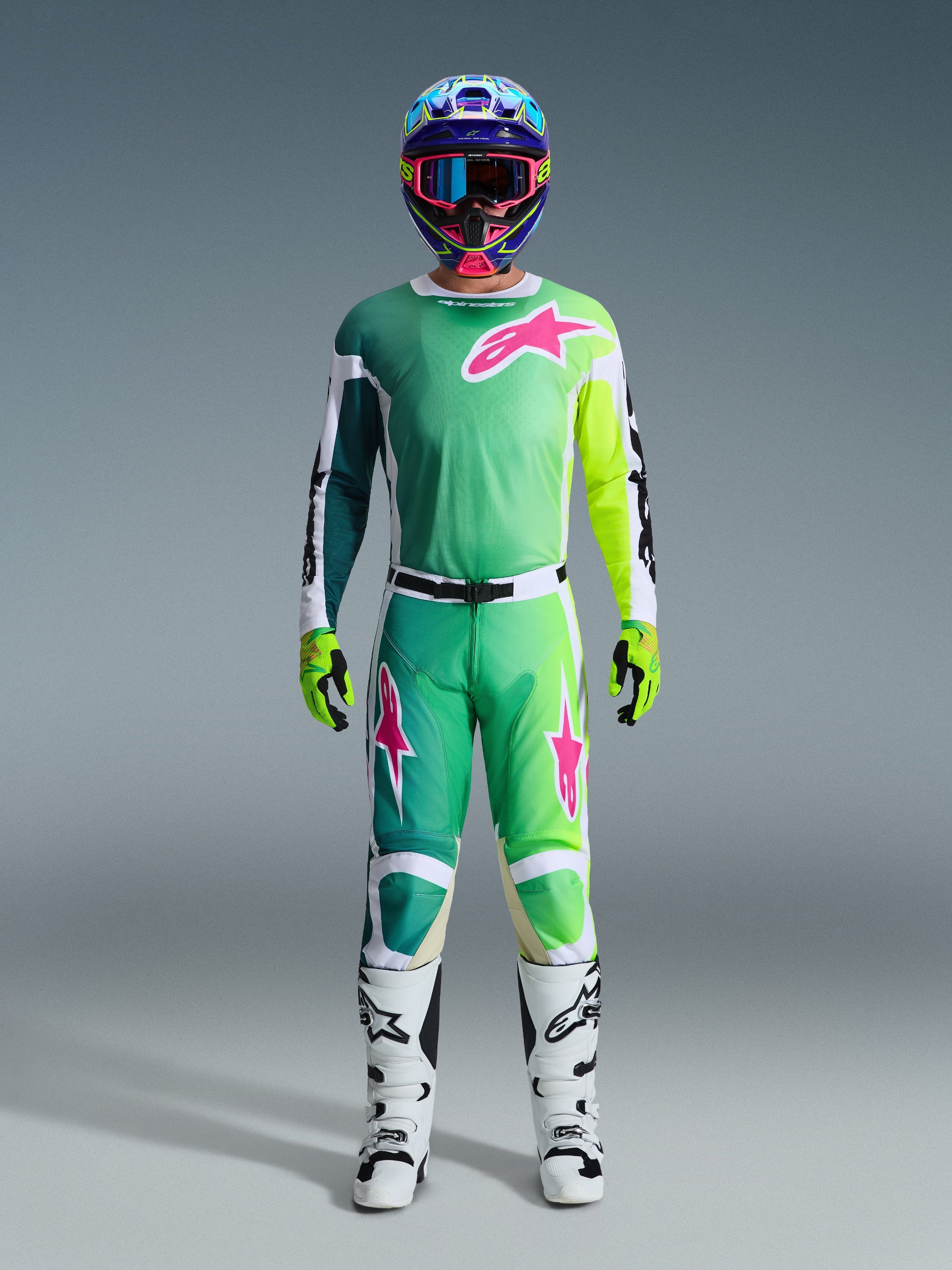 Alpinestars 2026 Racer Portl Gear Set, motocyklista motocrossowy w pełnym stroju wyścigowym, zielona bluza i spodnie z różowymi i białymi grafikami, niebieski wielokolorowy kask, biało-czarne buty i zielone rękawice, dopasowany zestaw do wyścigów terenowych