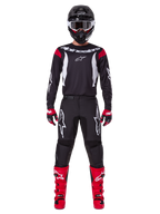 Alpinestars 2025 Fluid Haul Gear Set, zawodnik motocrossowy w pełnym stroju wyścigowym, czarna koszulka z czerwonymi akcentami na ramionach i białym logo, czarne spodnie z białym logo gwiazdy, jaskrawoczerwone buty, czerwono-czarne rękawice, czarny kask z białymi goglami, spójny wyczynowy styl off-road