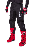 Alpinestars 2025 Fluid Haul Pants, spodnie motocrossowe, czarne z dużym białym logo Alpinestars, noszone z jaskrawoczerwonymi butami i czerwono-czarnymi rękawicami, wytrzymała konstrukcja z poliestru 600D o ergonomicznym kroju do jazdy terenowej