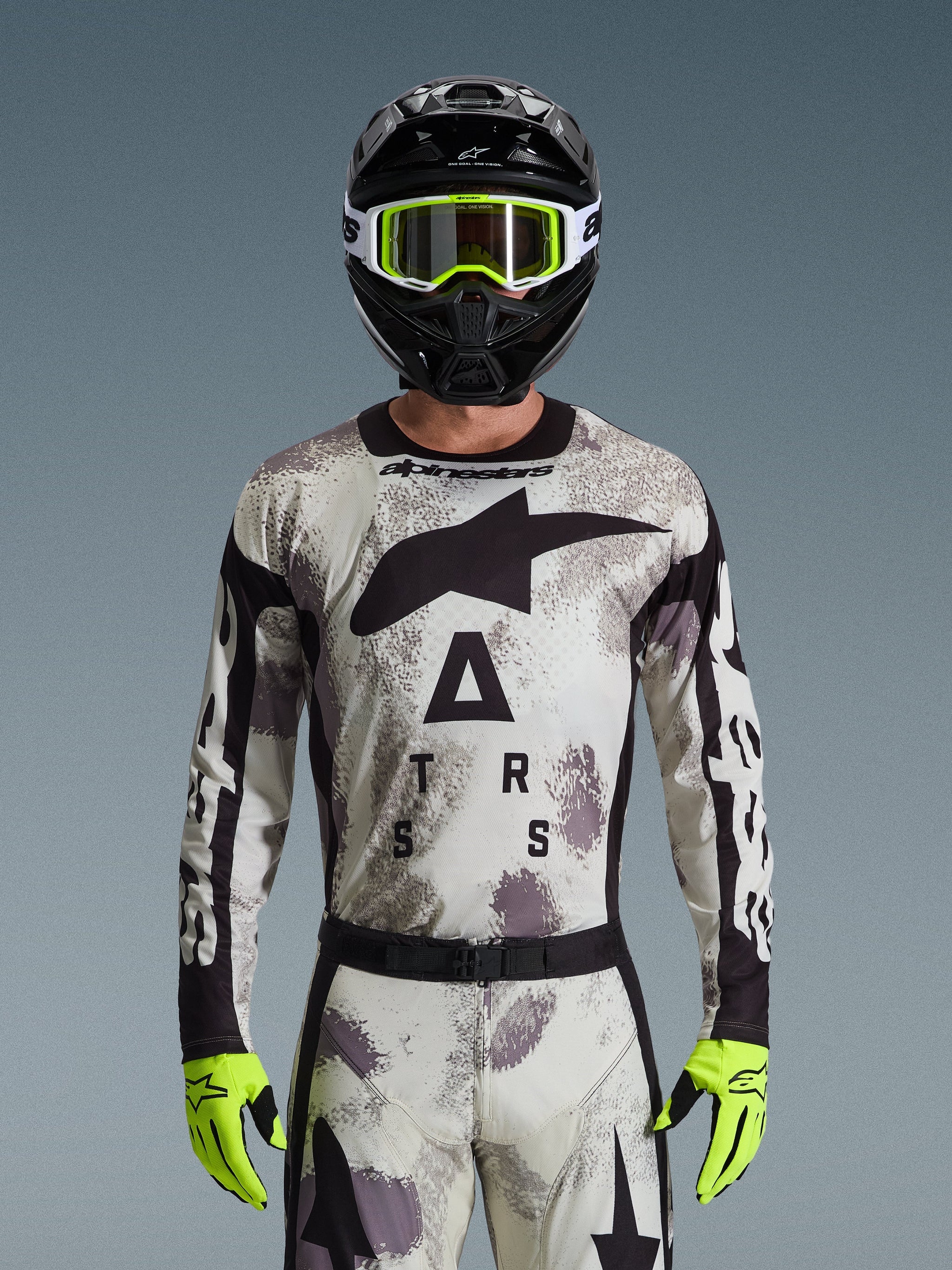 Motocyklista motocrossowy w koszulce Alpinestars 2026 Racer Lahnd, wzór Sand Camo w odcieniach szarości i bieli, duże czarne logo Astar oraz branding na klatce piersiowej, czarny kask, neonowożółte gogle i rękawice, profesjonalny zestaw do jazdy off-road