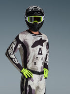 Motocyklista motocrossowy ubrany w koszulkę i zestaw Alpinestars 2026 Racer Lahnd w kolorze Sand Camo, jasnoszary wzór kamuflażu z czarnym logo, czarny kask z biało-neonowożółtymi goglami, jaskrawe neonowożółte rękawice, profesjonalna odzież wyścigowa off-road