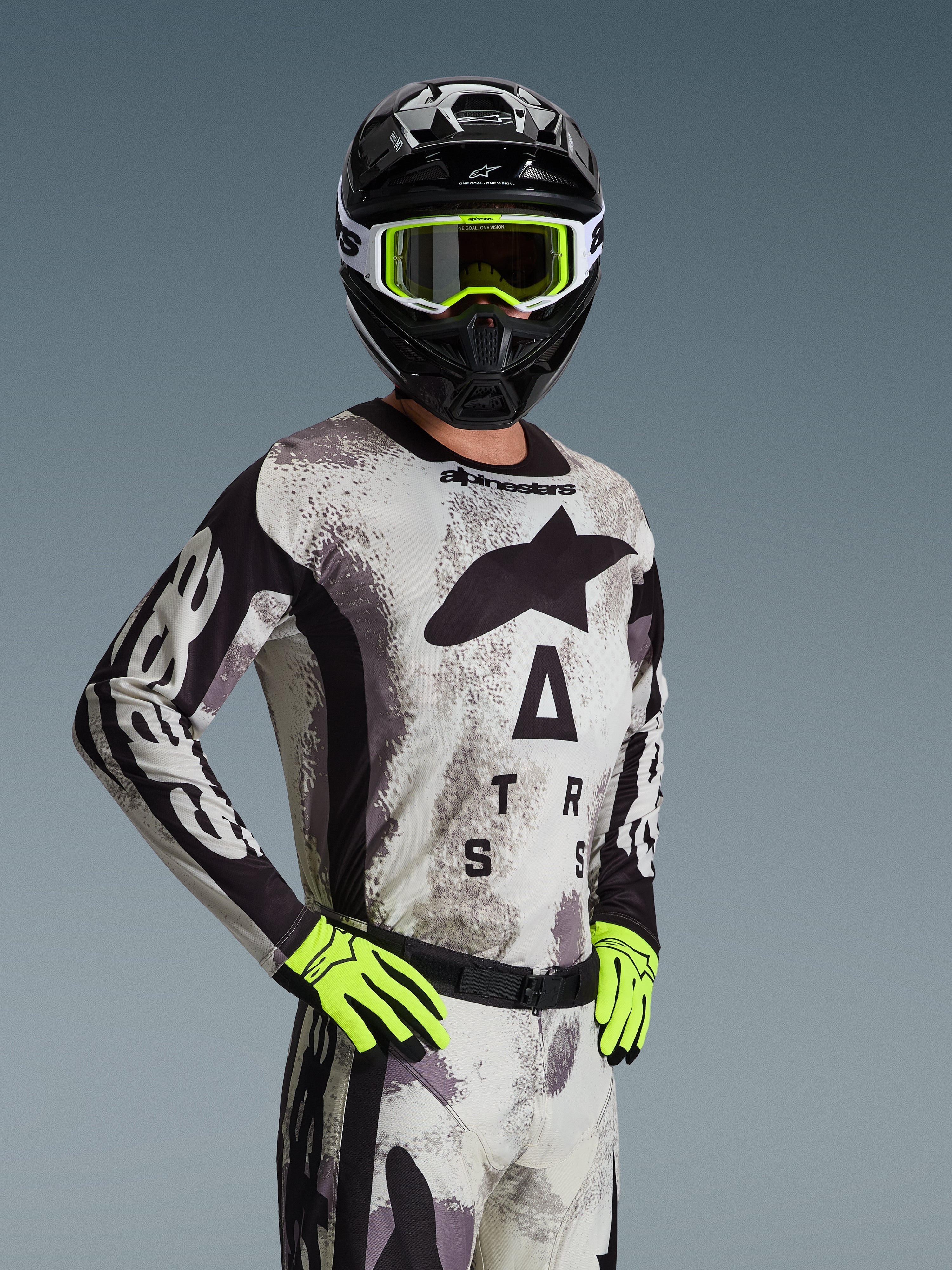 Motocyklista motocrossowy ubrany w koszulkę i zestaw Alpinestars 2026 Racer Lahnd w kolorze Sand Camo, jasnoszary wzór kamuflażu z czarnym logo, czarny kask z biało-neonowożółtymi goglami, jaskrawe neonowożółte rękawice, profesjonalna odzież wyścigowa off-road