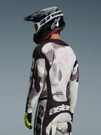 Zawodnik motocrossowy od tyłu w zestawie Alpinestars 2026 Racer Lahnd, koszulka i spodnie w szaro-białym kamuflażu Sand Camo z czarnymi akcentami, czarny kask z białym logo Alpinestars, neonowożółte i czarne rękawice, profesjonalny strój off-road