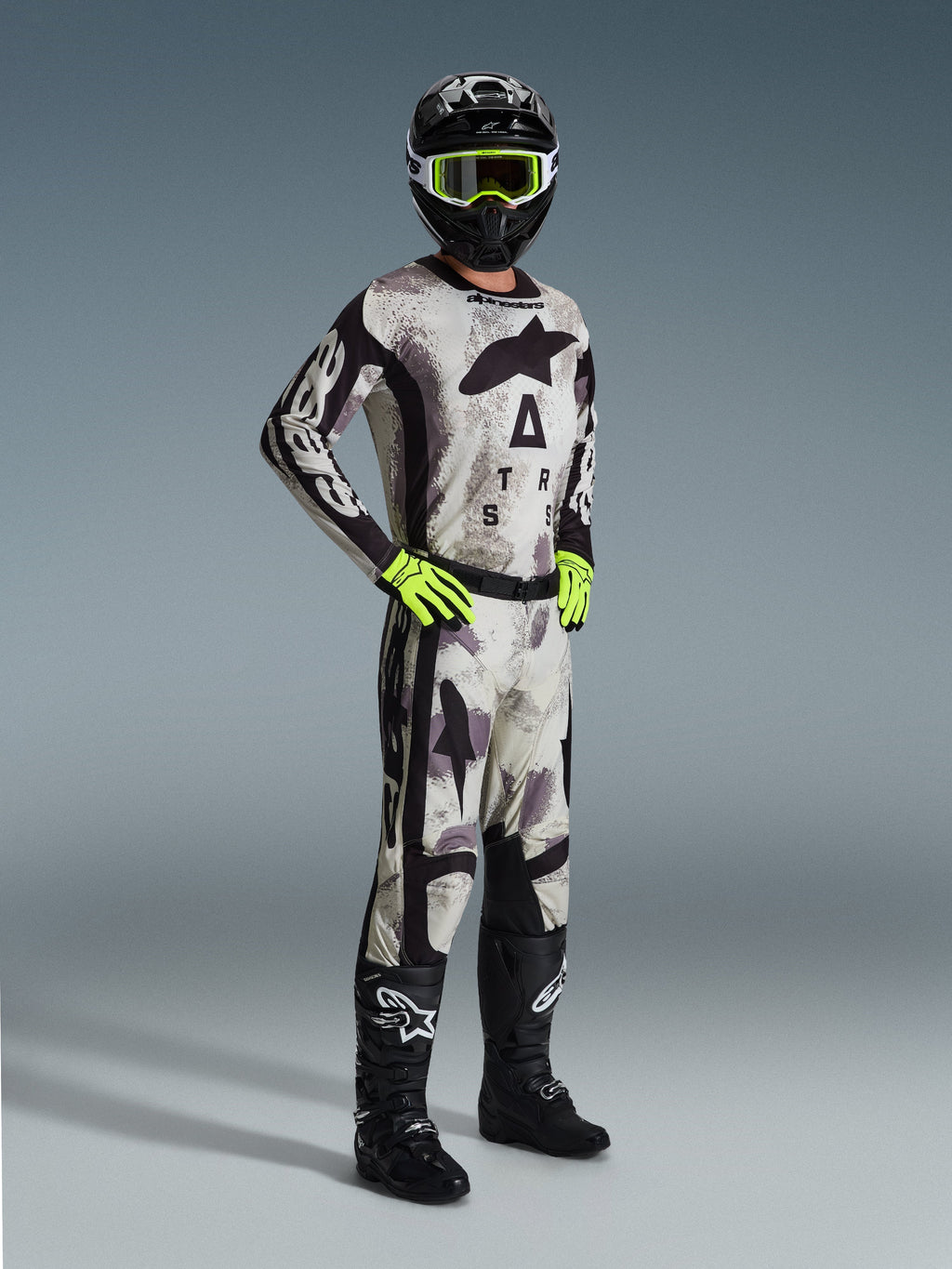 Zawodnik motocrossowy w pełnym stroju wyścigowym Alpinestars, spodnie Racer Lahnd 2026 w kolorze Sand Camo, dopasowana koszulka camo z wyraźnym brandingiem, czarny kask z neonowo-żółtymi goglami, neonowo-zielone rękawice, czarne buty, profesjonalny zestaw do jazdy off-road