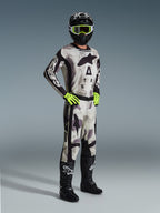 Zawodnik motocrossowy w pełnym stroju wyścigowym Alpinestars, spodnie Racer Lahnd 2026 w kolorze Sand Camo, dopasowana koszulka camo z wyraźnym brandingiem, czarny kask z neonowo-żółtymi goglami, neonowo-zielone rękawice, czarne buty, profesjonalny zestaw do jazdy off-road