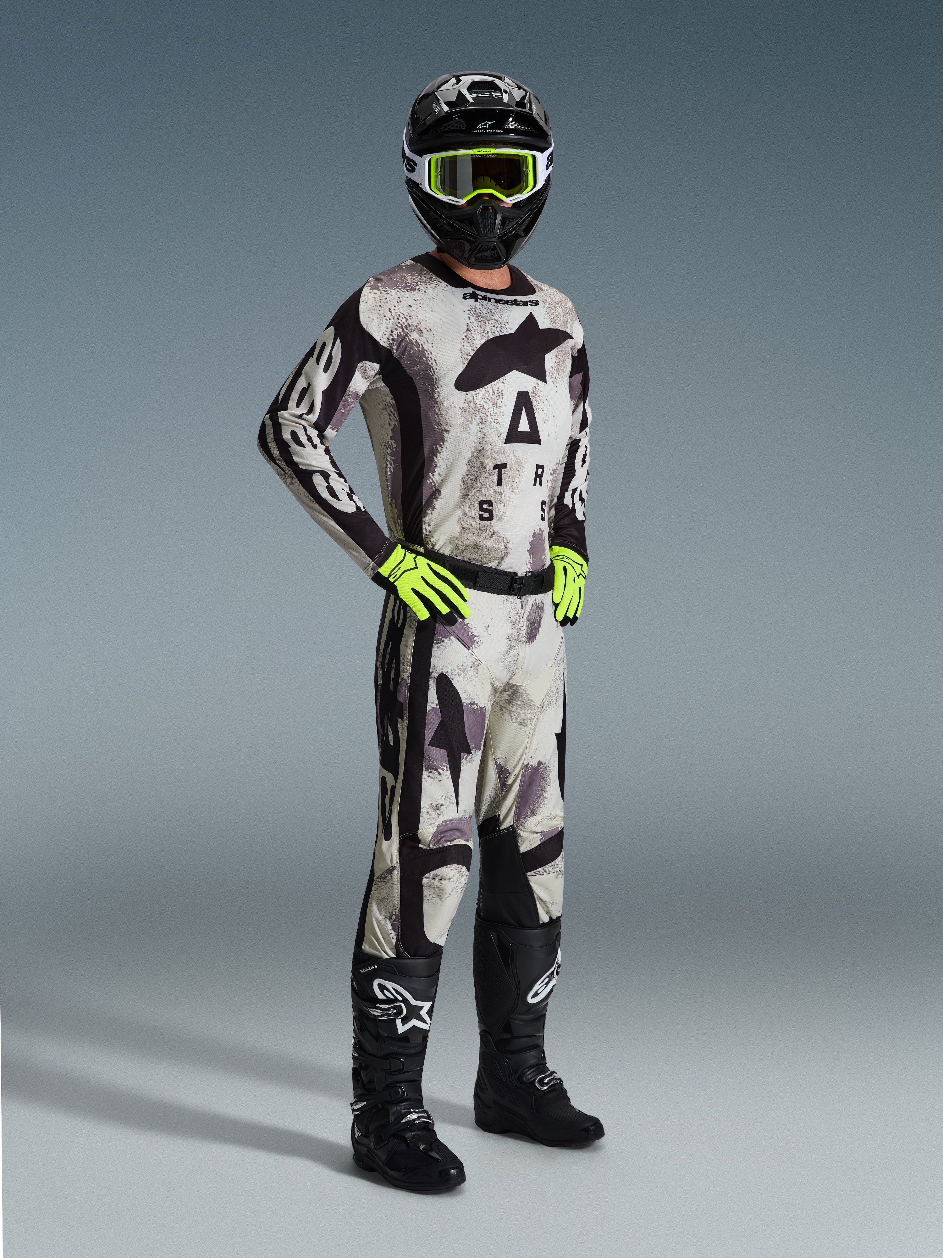Zawodnik motocrossowy w pełnym stroju wyścigowym Alpinestars, spodnie Racer Lahnd 2026 w kolorze Sand Camo, dopasowana koszulka camo z wyraźnym brandingiem, czarny kask z neonowo-żółtymi goglami, neonowo-zielone rękawice, czarne buty, profesjonalny zestaw do jazdy off-road