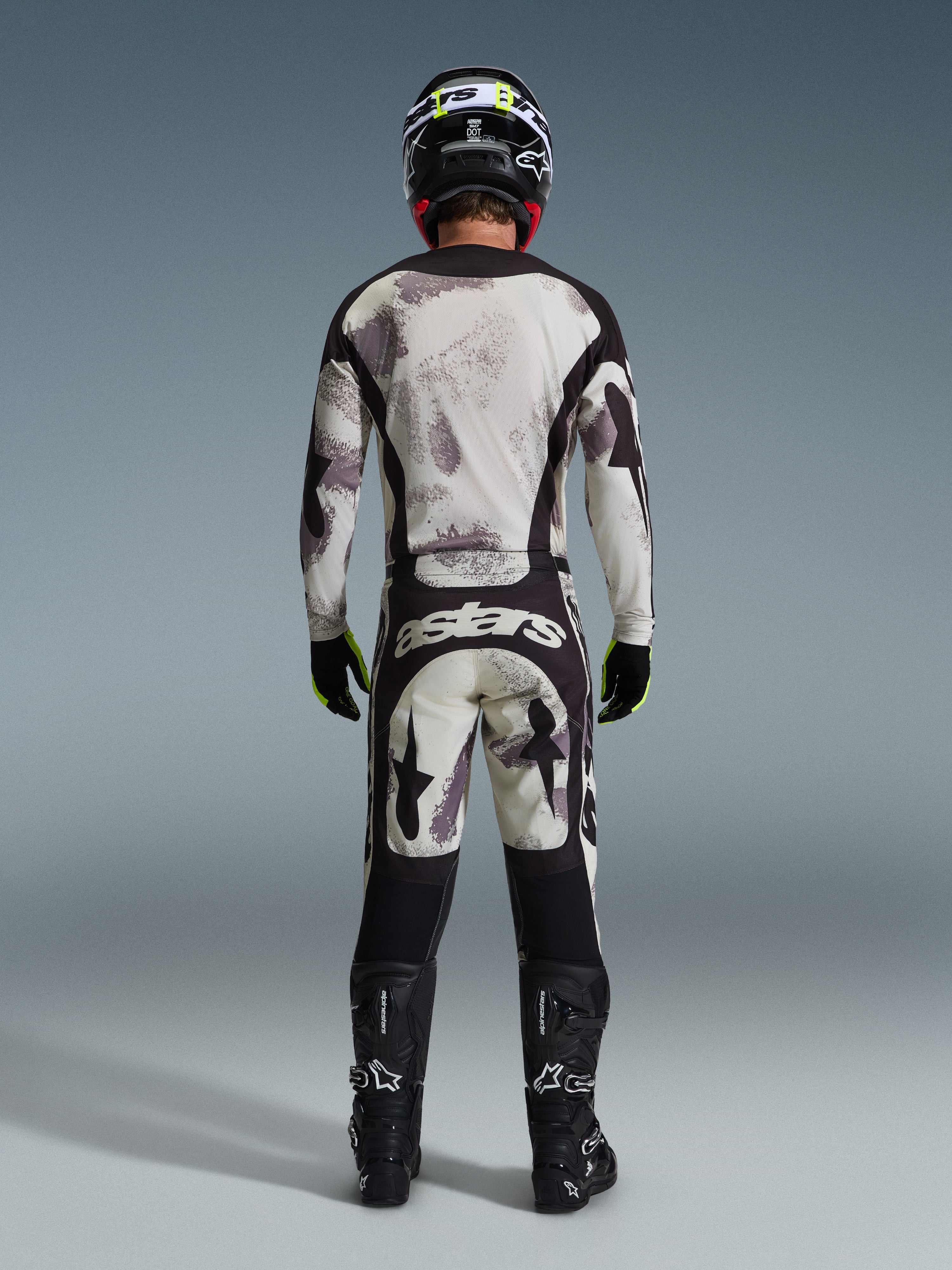 Zawodnik motocrossowy w kompletnym zestawie Alpinestars 2026 Racer Lahnd, widok z tyłu, bluza i spodnie Sand Camo z jasnoszarym i czarnym abstrakcyjnym wzorem, czarny kask, czarno-neonowożółte rękawice, czarne buty, logo „astars” na tyle spodni